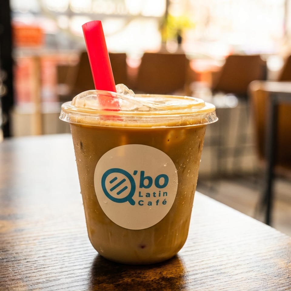 Iced coffee in a clear cup with a red straw and Qbo Latin Café logo on a wooden café table.