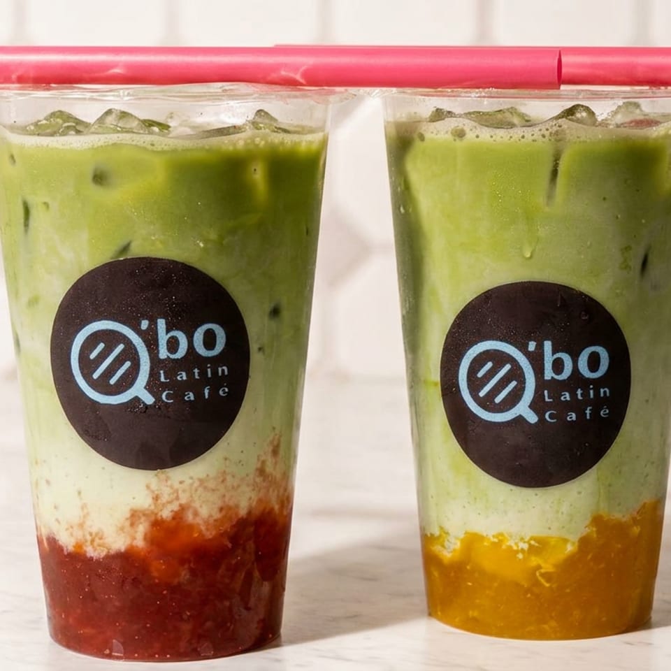 Two iced green matcha drinks with colorful fruit layers in branded Q’bo Latin Café cups and red straws.
