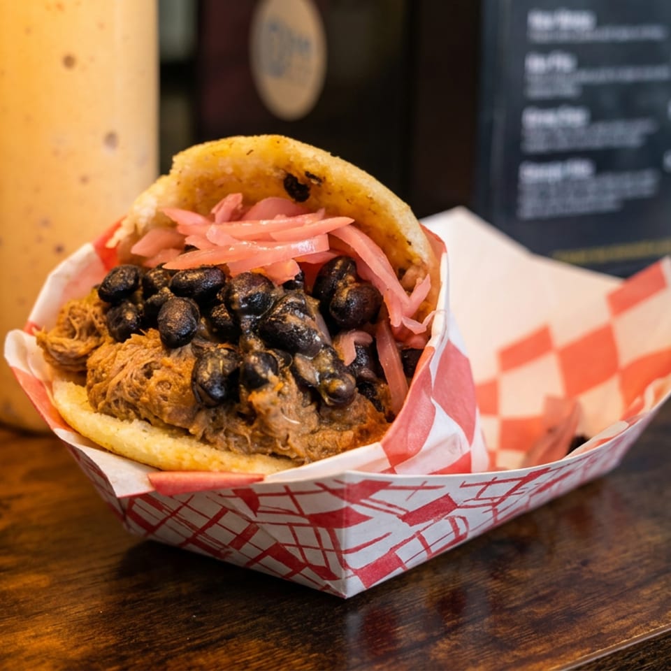 Loaded sandwich with crispy fried filling, black beans, and pickled onions in a red-and-white paper tray on a wooden counter.