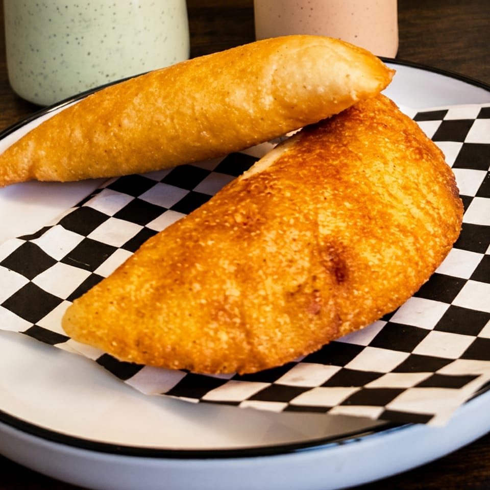 Two golden fried empanadas rest on a black-and-white checkered liner atop a white plate on a wooden table.