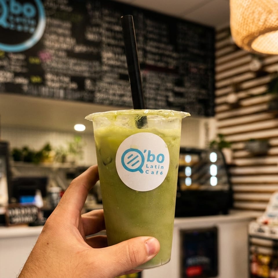 Hand holds a green iced drink with a straw and Ybo Latin Café logo inside a warmly lit café.