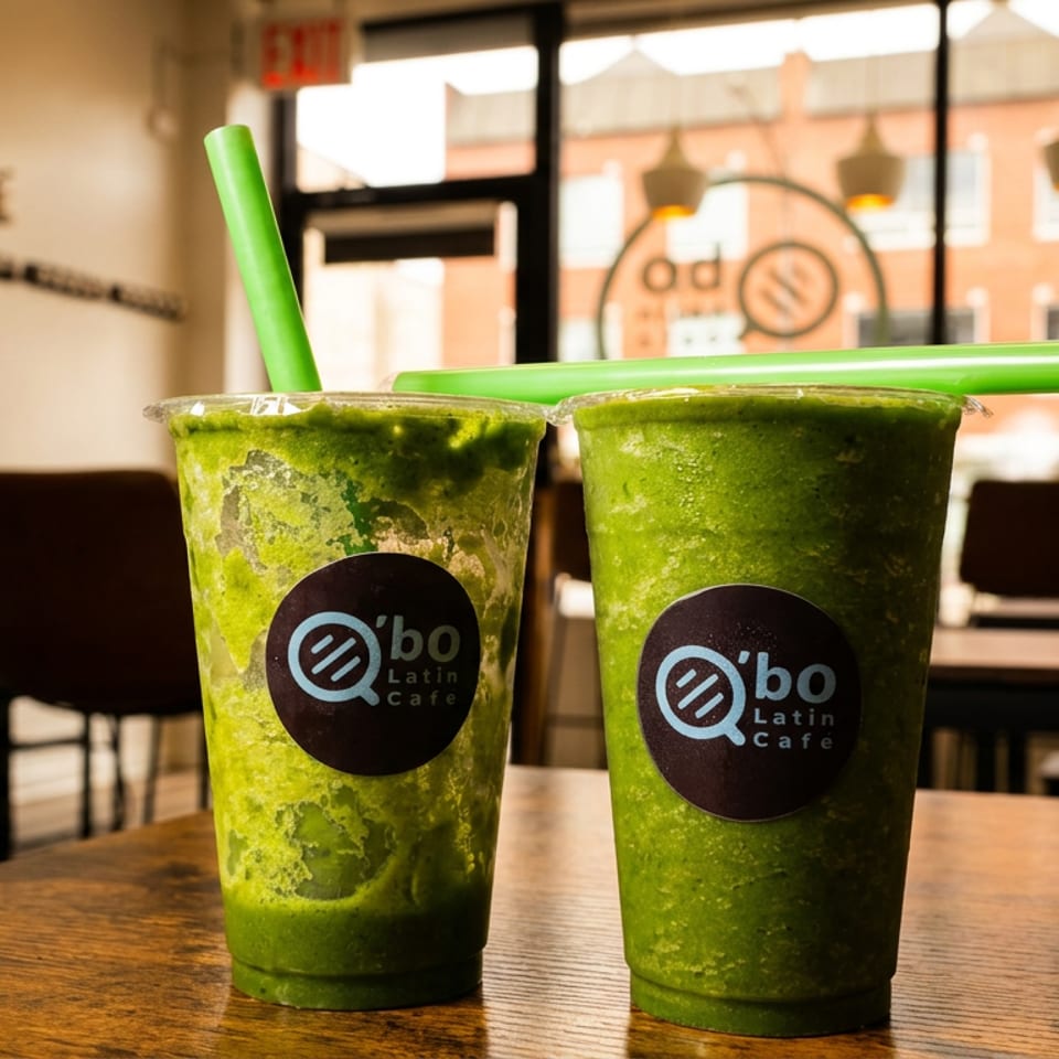 Two green smoothies with branded straws sit on a wooden table inside Qbo Latin Café.